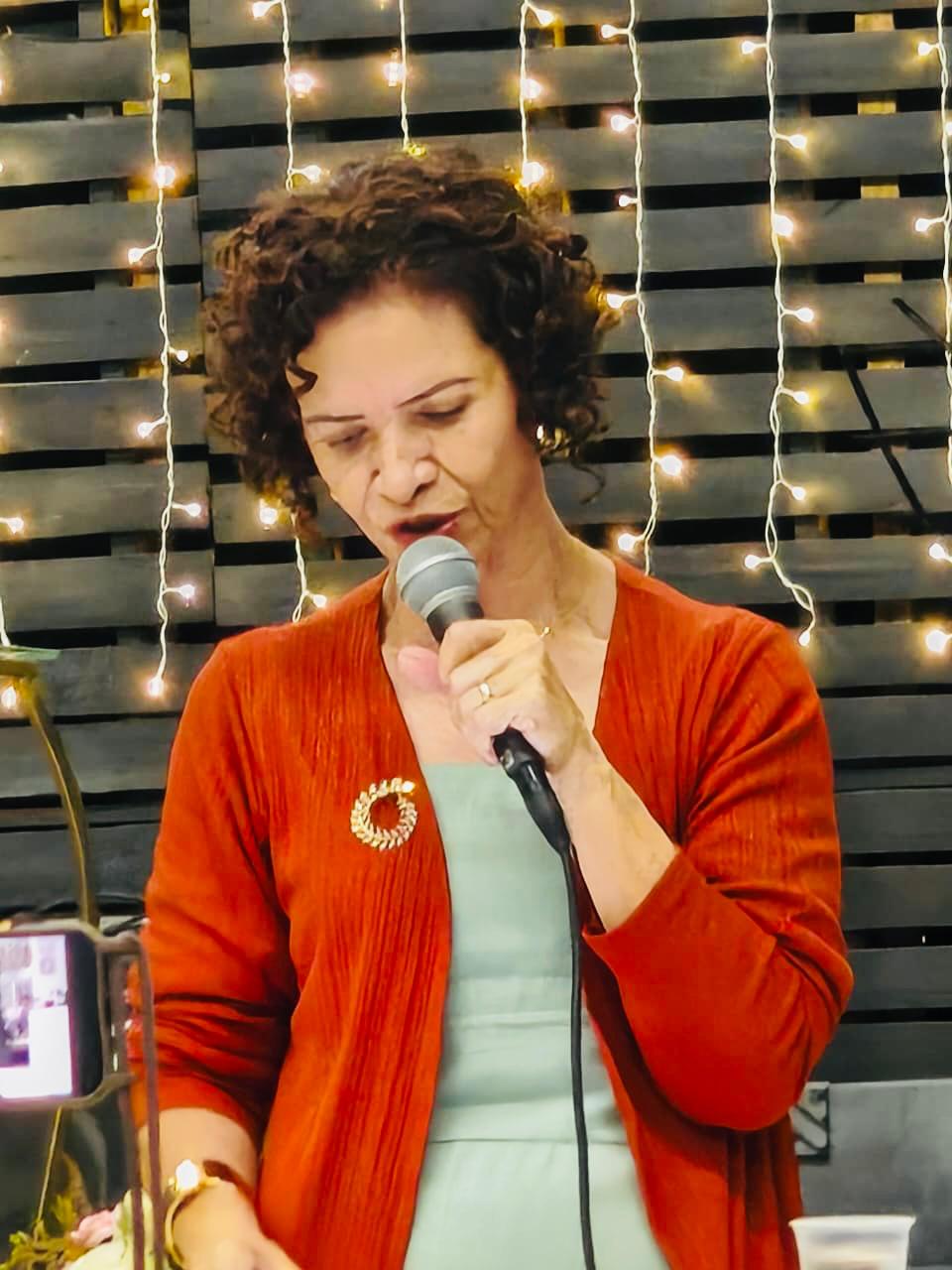 Janethi Menezes falando ao microfone em um evento, de casaco laranja, diante de uma parede com luzes